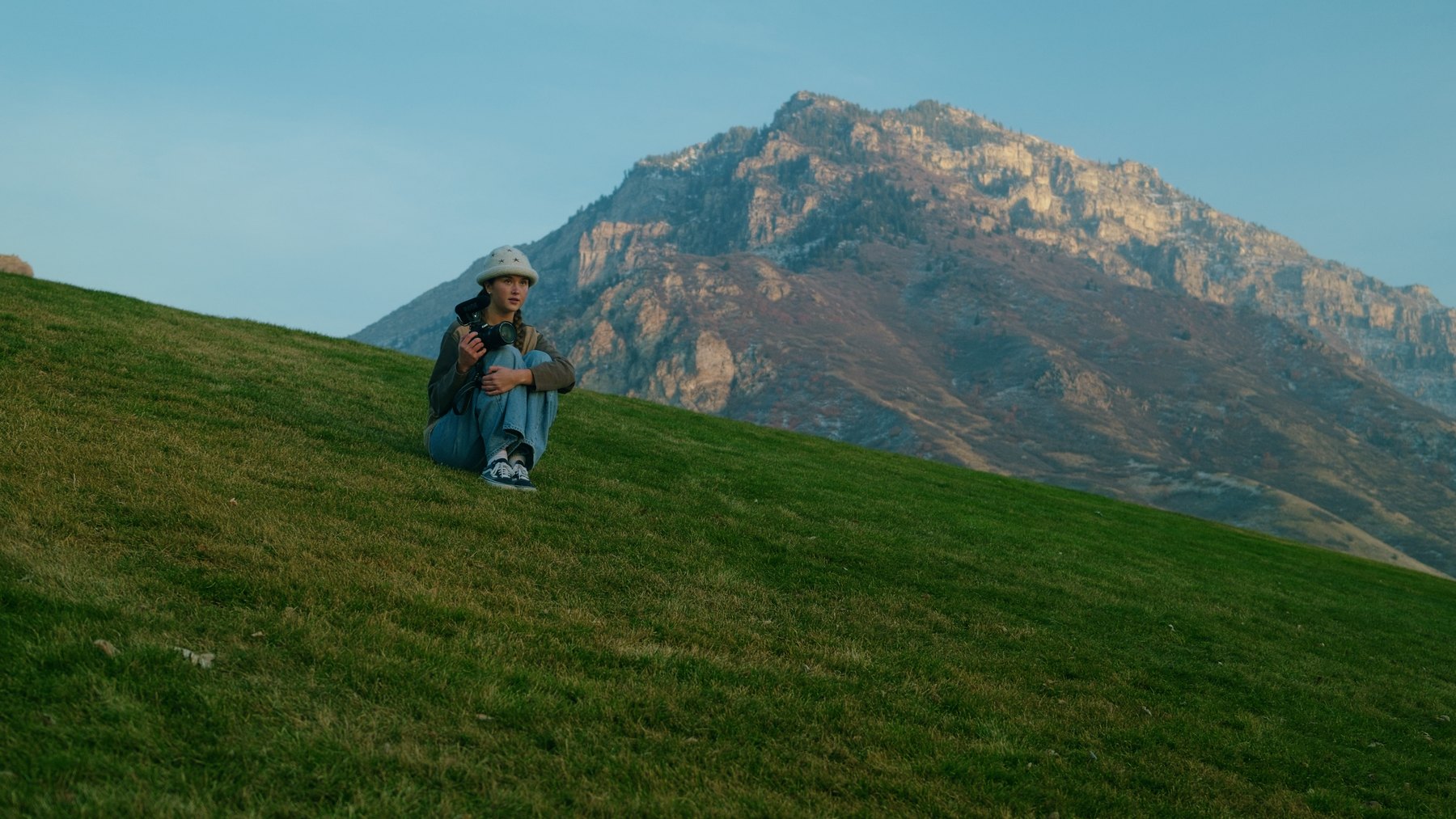 Bailey Nye sitting on a grassy hillside below a mountain
