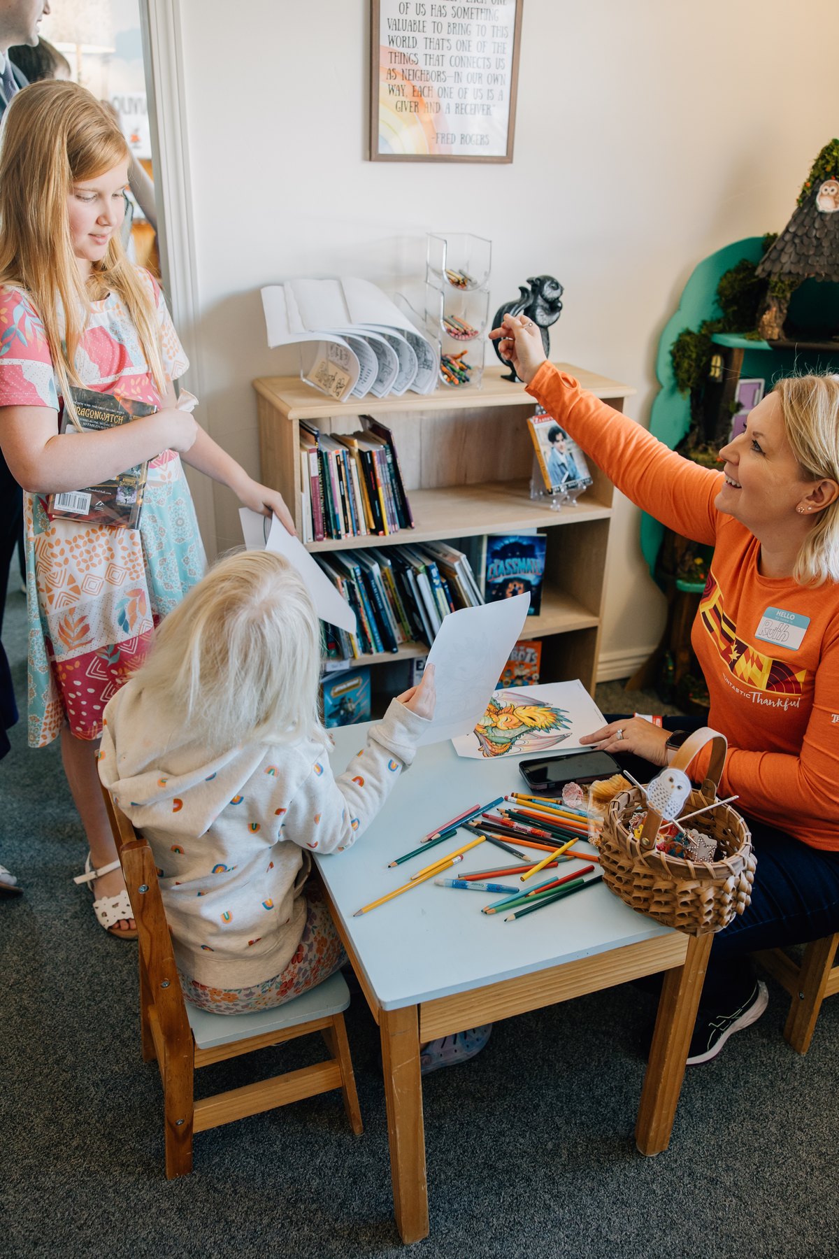 A volunteer handing colouring pages to two children at a small table