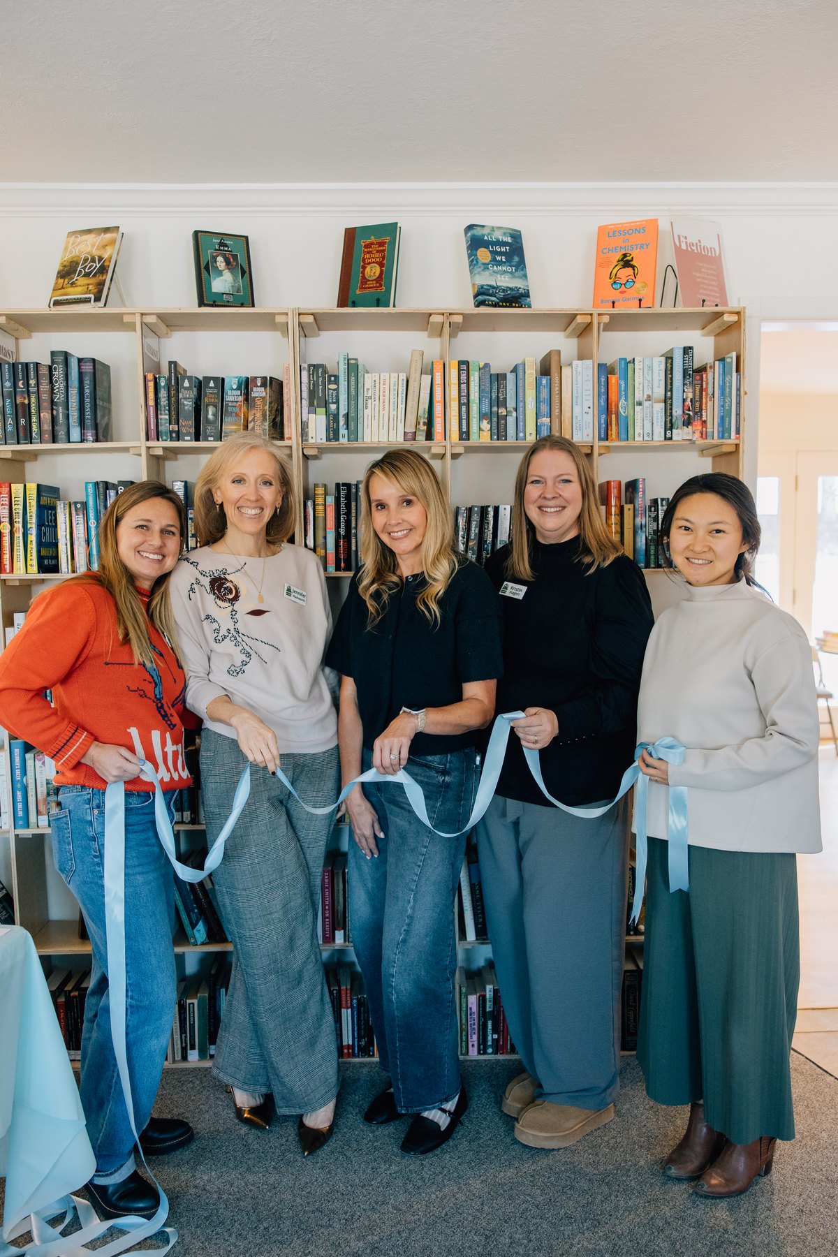 Five founders of The Friends of Alpine Library holding a blue ribbon in front of the fiction shelves